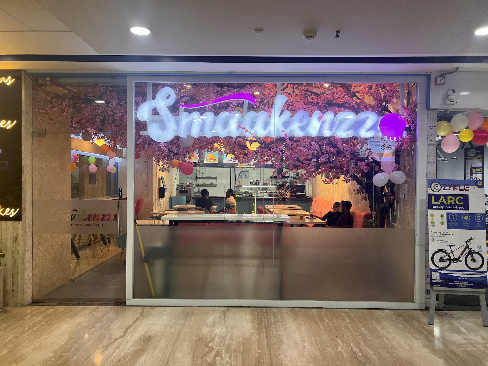 The Smaakenzzo storefront inside Preston Prime Mall, the name in white neon script against blossom branches, balloons at the door