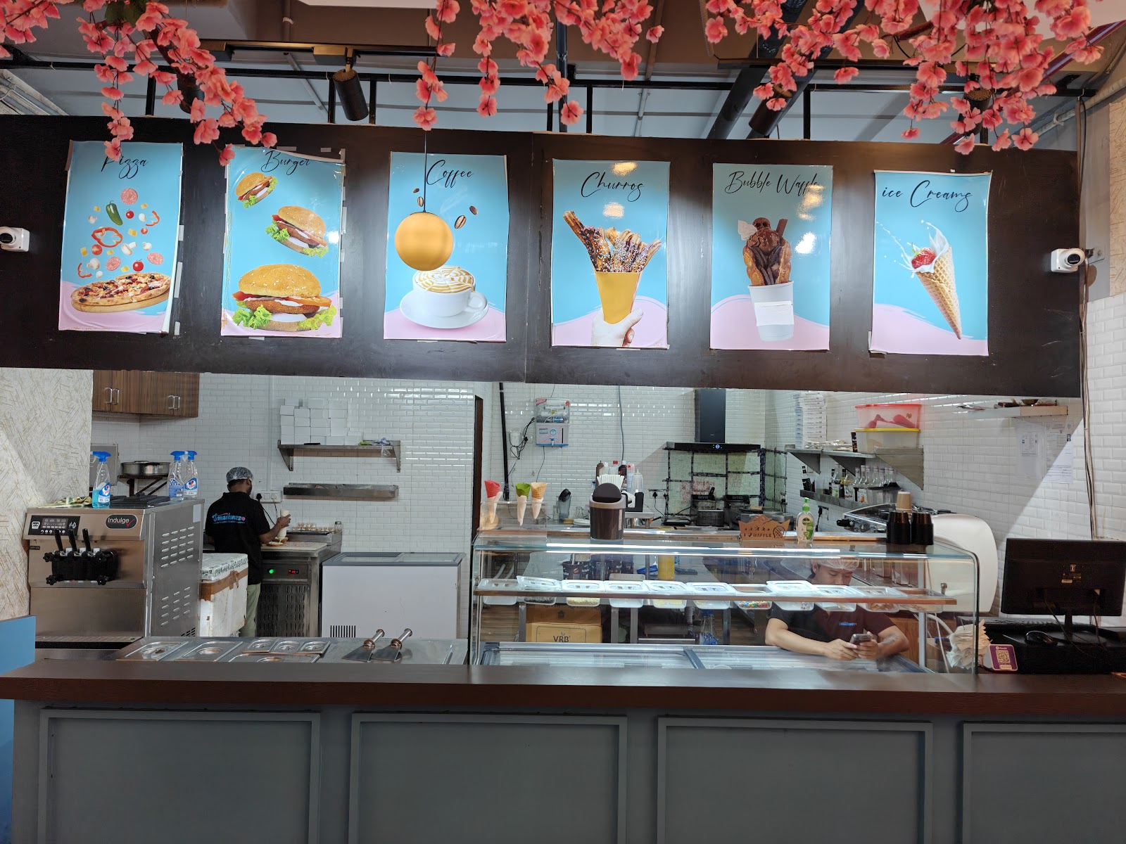 The service counter, with six lit menu boards above it reading Pizza, Burger, Coffee, Churros, Bubble Waffle and Ice Creams, an ice cream cabinet below and staff at work