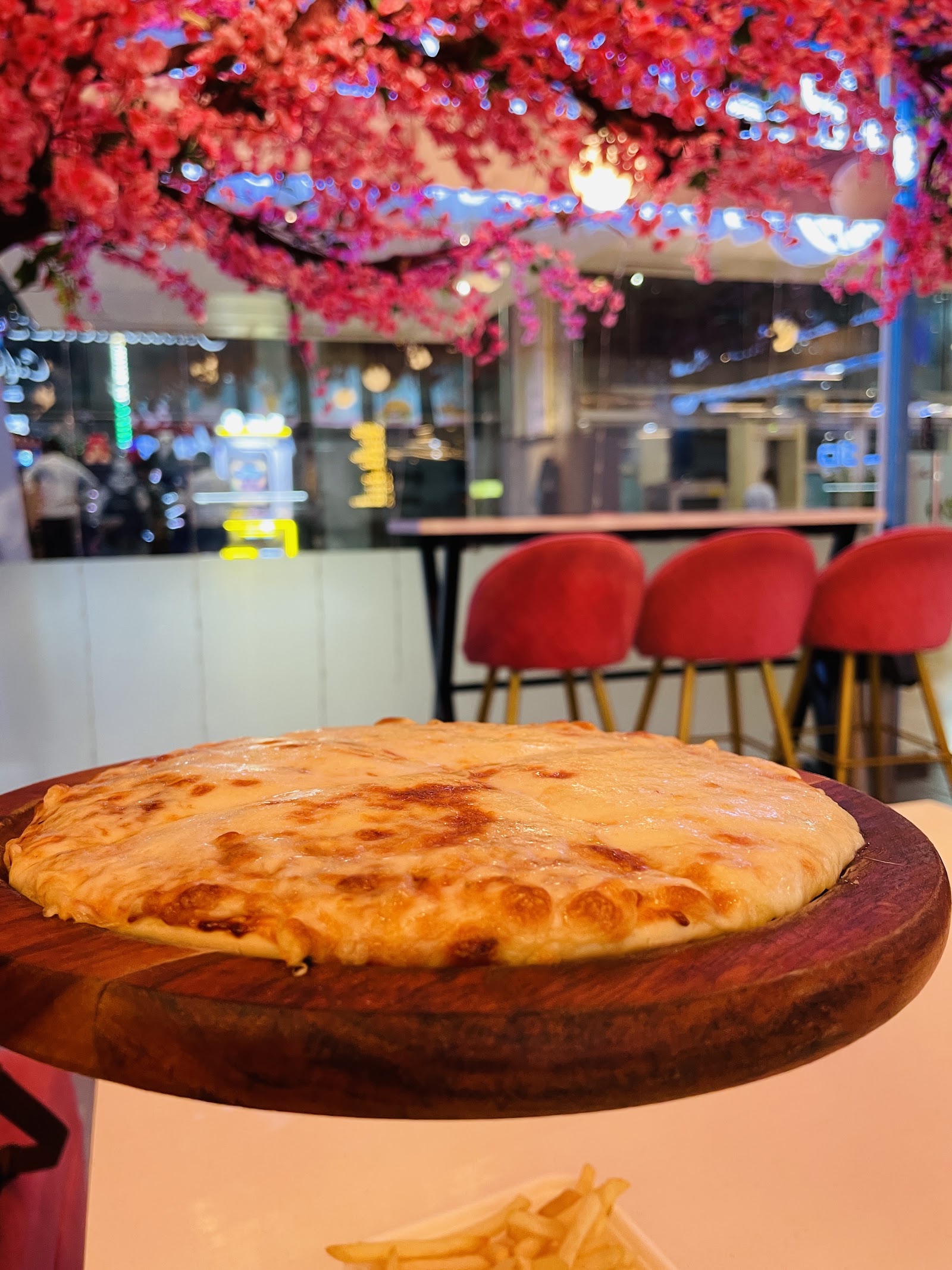 A cheese pizza on a round wooden board, blossom branches and red velvet bar stools behind