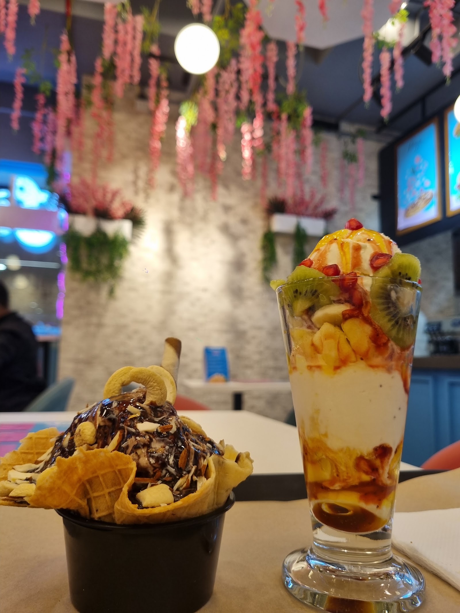 A chocolate sundae in a waffle basket beside a layered fruit parfait, under hanging pink blossom
